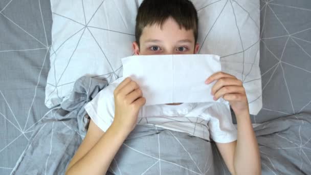 Мальчик с гриппом и лихорадкой лежит в постели и высморкается бумажной салфеткой, концепция сезонных вирусных заболеваний — стоковое видео