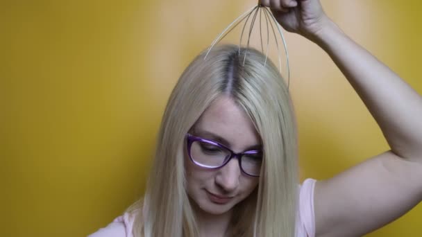 Uma mulher loira caucasiana usando óculos desfrutar de cabeça, massagem no couro cabeludo por anti estresse acupuntura ferramenta de polvo de metal, equipamento, ela fecha os olhos com prazer, fundo amarelo — Vídeo de Stock