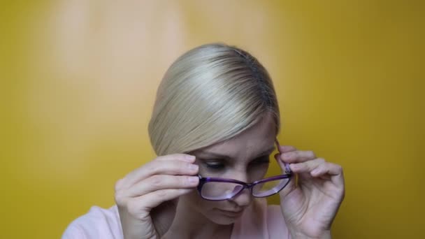 Een blonde vrouw van middelbare leeftijd zet stijlvolle modieuze glazen op en glimlacht naar de camera tegen gele achtergrond, bijziendheid, astigmatisme en oogheelkundige ziekten. — Stockvideo