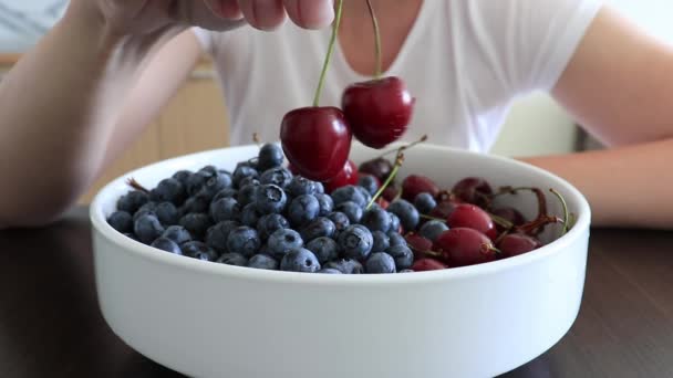 Mujer sosteniendo cereza dulce por el tallo y retorciéndolo debajo del tazón, plato lleno de bayas jugosas y sabrosas en la cocina, vista de cerca — Vídeo de stock