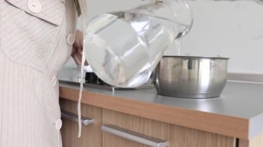 Sürahiden metal bir tencereye saf içme suyu döken bir kadın, mutfaktaki sürahi ve kaynatmak için ocağa koyan bir kadın..