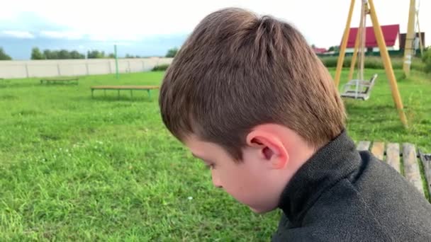 Хлопчик сидить на лавці на дитячому майданчику, дивлячись в камеру і роблячи смішні обличчя, використовуючи брови — стокове відео