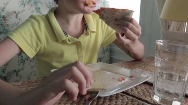 Un chico comiendo codiciosamente pizza en un restaurante, está sosteniendo un pedazo de pizza, mordiéndola y masticándola — Vídeos de Stock