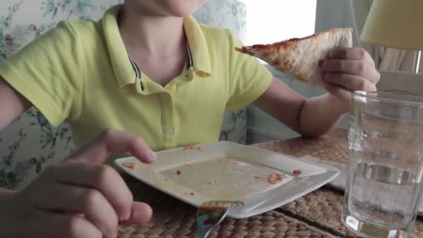 Ένα αγόρι άπληστα τρώει πίτσα σε ένα εστιατόριο, κρατάει ένα κομμάτι πίτσα, το δαγκώνει και μασάει — Αρχείο Βίντεο