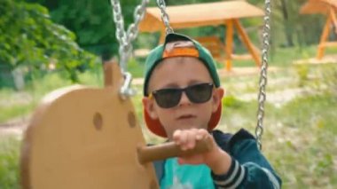 Güneş ışığı altında bir parkta bir at şeklinde ahşap bir salıncak üzerinde boy mutlu gülümseyerek. Güneş gözlüğü ve bir kap parkta bir salıncak üzerinde eğleniyor