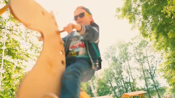 Joyeux garçon souriant sur une balançoire en bois en forme de cheval dans un parc à la lumière du soleil. Un enfant en lunettes de soleil et une casquette s'amuse sur une balançoire dans le parc — Video