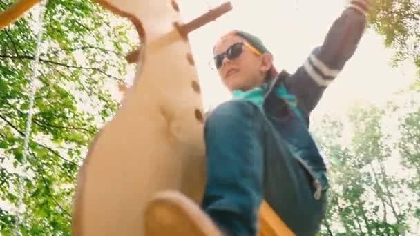 Szczęśliwy uśmiechający się chłopak na drewnianej huśtawce w formie konia w parku w słońcu. Dziecko w okulary przeciwsłoneczne i czapka jest zabawy na huśtawce w parku — Wideo stockowe