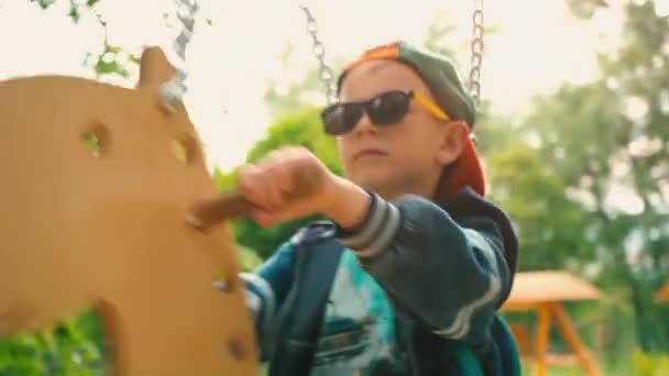 Szczęśliwy uśmiechający się chłopak na drewnianej huśtawce w formie konia w parku w słońcu. Dziecko w okulary przeciwsłoneczne i czapka jest zabawy na huśtawce w parku — Wideo stockowe