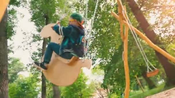 Happy smiling boy on a wooden swing in the form of a horse in a park in the sunlight. A child in sunglasses and a cap is having fun on a swing in the park — Stock Video