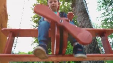 Güneş ışığı altında bir parkta bir uçak şeklinde ahşap bir salıncak üzerinde boy mutlu gülümseyerek. Bir çocuk bir salıncakta eğleniyor