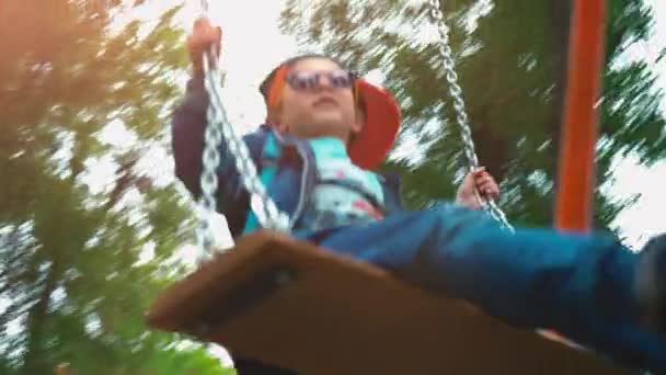 Petit garçon aux lunettes de soleil et coiffe verte balançant sur une balançoire, un enfant de 5 ans s'amuse sur une balançoire d'enfants entourée d'arbres verts — Video
