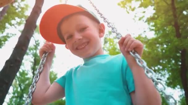 Sonriente niño de 5 años con una camiseta azul y con una gorra en la cabeza balanceándose en un columpio con cadenas de acero, un niño se divierte en un columpio para niños rodeado de árboles verdes — Vídeo de stock