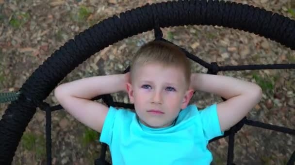 Le garçon de cinq ans aux yeux bleus est allongé sur une balançoire ronde, lève les mains derrière la tête, regarde le ciel et les rêves, l'enfant se repose, couché sur la balançoire — Video