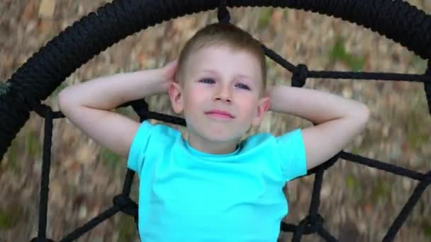 The blue-eyed five-year-old boy in blue t-shirt lies on a round swing, throws up his hands behind his head, looks up at the sky and dreams, the child is resting, lying on the swing — Stock Video