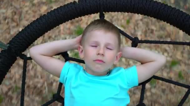 Um menino de 5 anos de olhos azuis com uma camiseta azul sonha com os olhos fechados, deitado num baloiço redondo. Uma criança sorri enquanto pensa em um agradável pairando sobre o chão em um balanço — Vídeo de Stock