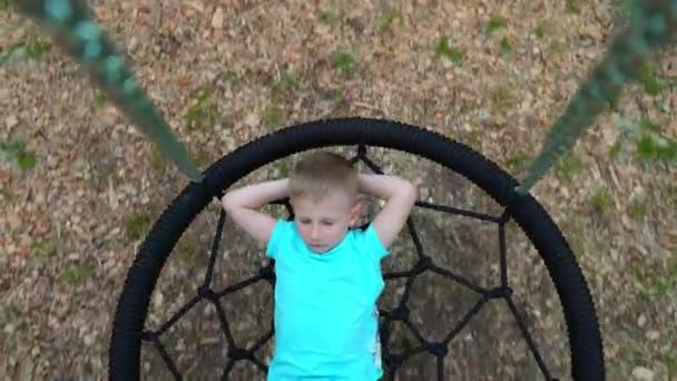 A blue-eyed, 5-year-old boy in a blue T-shirt dreams with his eyes closed, lying on a round swing. A child smiles while thinking about a pleasant hovering above the ground on a swing — Stock Video