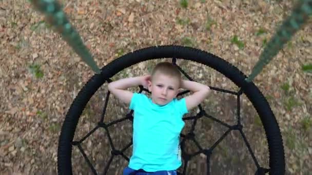 The blue-eyed five-year-old boy in blue t-shirt lies on a round swing with his hands behind his head, looks at the camera and smiles, the child is resting, lying on the swing — Stock Video