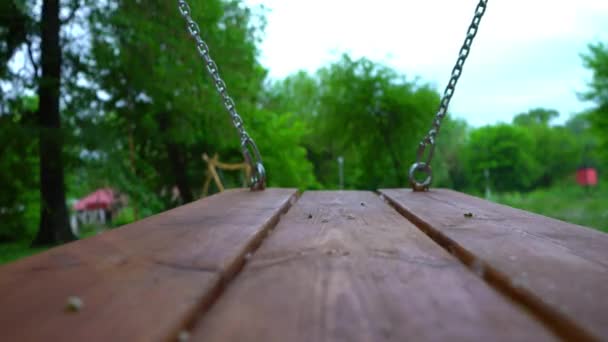 Holzschaukel an Stahlketten schaukelt in einem leeren Kinderpark — Stockvideo