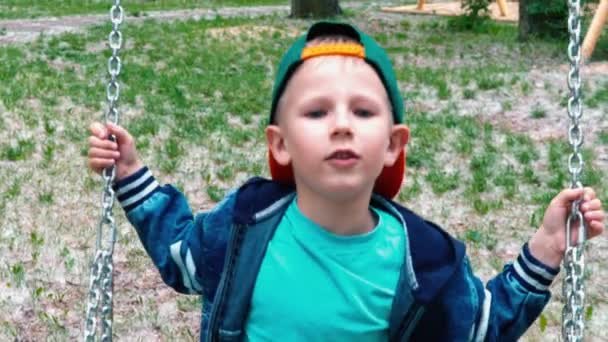 El niño de 5 años se rompe del columpio, pero no cae al suelo, el niño con una camiseta azul y una gorra verde no descansa en un columpio en el parque verde, en cámara lenta — Vídeos de Stock