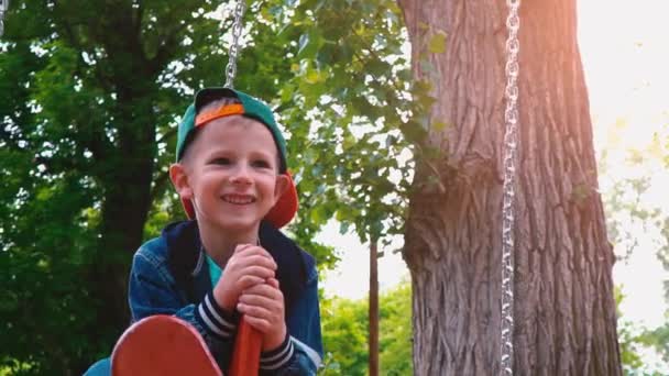 Joyeux garçon souriant sur une balançoire en bois sous la forme d'un avion dans un parc à la lumière du soleil. Un enfant s'amuse sur une balançoire au ralenti — Video