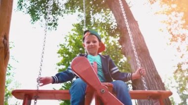 Güneş ışığı altında bir parkta bir uçak şeklinde ahşap bir salıncak üzerinde boy mutlu gülümseyerek. Bir çocuk bir salıncak, yavaş hareket eğleniyor