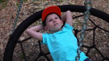 Beş yaşında mavi gözlü çocuk t-shirt yuvarlak salıncakta yatıyor, ellerini başının arkasına kadar atar, gökyüzü ve rüyalar baktı mavili çocuğa çocuk, salıncak, ağır çekim yalan dinleniyor