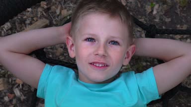 Beş yaşında mavi gözlü çocuk t-shirt yuvarlak salıncakta yatıyor, ellerini başının arkasına kadar atar, gökyüzü ve rüyalar baktı mavili çocuğa çocuk, salıncak, ağır çekim yalan dinleniyor