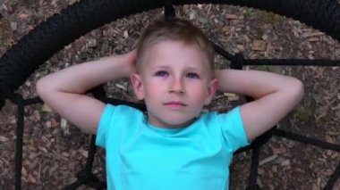 Beş yaşında mavi gözlü çocuk t-shirt yuvarlak salıncakta yatıyor, ellerini başının arkasına kadar atar, gökyüzü ve rüyalar baktı mavili çocuğa çocuk, salıncak, ağır çekim yalan dinleniyor