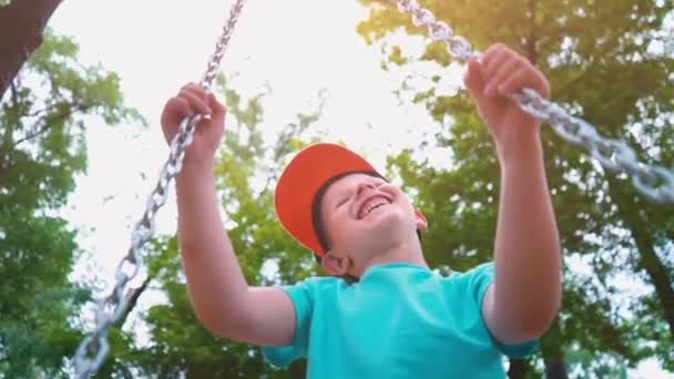 Sonriente niño de 5 años con una camiseta azul y con una gorra en la cabeza balanceándose en un columpio con cadenas de acero, un niño se divierte en un columpio para niños rodeado de árboles verdes, cámara lenta — Vídeo de stock