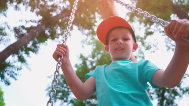 Sonriente niño de 5 años con una camiseta azul y con una gorra en la cabeza balanceándose en un columpio con cadenas de acero, un niño se divierte en un columpio para niños rodeado de árboles verdes, cámara lenta — Vídeo de stock