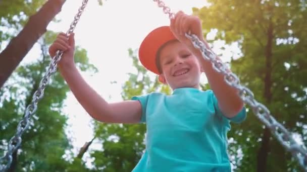 Sonriente niño de 5 años con una camiseta azul y con una gorra en la cabeza balanceándose en un columpio con cadenas de acero, un niño se divierte en un columpio para niños rodeado de árboles verdes, cámara lenta — Vídeos de Stock