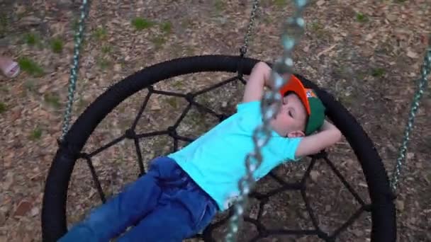 Голубоглазый пятилетний мальчик в голубой футболке лежит на качелях, поднимает руки за голову, смотрит на небо и видит сны, ребенок отдыхает, лежит на качелях, медленное движение — стоковое видео