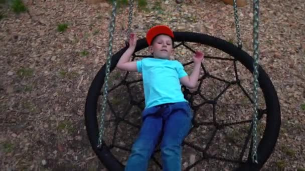 A criança de 5 anos em uma camiseta azul está em um balanço com correntes de ferro em um parque verde, balança em um balanço e sonha com algo, câmera lenta — Vídeo de Stock