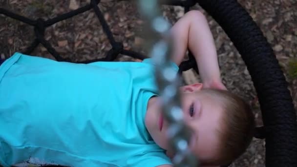 Il bambino di cinque anni dagli occhi azzurri con la maglietta blu giace su un'altalena rotonda, alza le mani dietro la testa, alza lo sguardo al cielo e sogna, il bambino riposa, sdraiato sull'altalena, al rallentatore — Video Stock