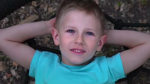 De blauwogige five-year-old boy blauw t-shirt ligt op een ronde schommel, gooit zijn handen achter zijn hoofd, kijkt omhoog naar de hemel en dromen, het kind is rust, liggend op de schommel, slow-motion — Stockvideo