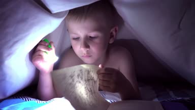 Bir çocuk geceleri bir el feneri ile battaniye altında bir kitap okur. ışık saç ve mavi gözlü bir çocuk