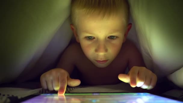 Young blond boy under covers playing on tablet a computer game. — Stock Video