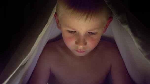 Young blond boy under covers playing on tablet a computer game. — Stock Video
