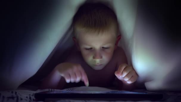 Kleiner blonder Junge spielt unter einer Decke auf dem Tablet ein Computerspiel. — Stockvideo