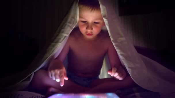 Young blond boy under covers playing on tablet a computer game. — Stock Video