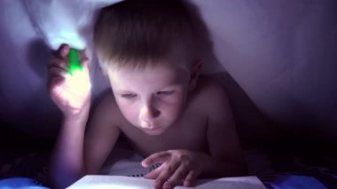 Bir çocuk geceleri bir el feneri ile battaniye altında bir kitap okur. ışık saç ve mavi gözlü bir çocuk