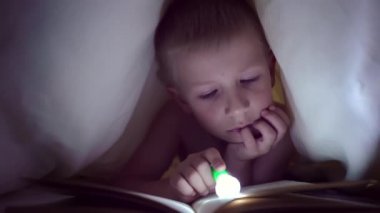 Bir çocuk geceleri bir el feneri ile battaniye altında bir kitap okur. ışık saç ve mavi gözlü çocuk. Yatmadan önce bir peri masalı