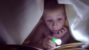 Bir çocuk geceleri bir el feneri ile battaniye altında bir kitap okur. ışık saç ve mavi gözlü bir çocuk