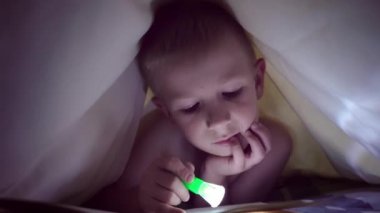 Bir çocuk geceleri bir el feneri ile battaniye altında bir kitap okur. ışık saç ve mavi gözlü bir çocuk