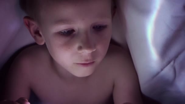 Un enfant lit un livre sous des couvertures avec une lampe de poche la nuit. garçon aux cheveux clairs et aux yeux bleus — Video