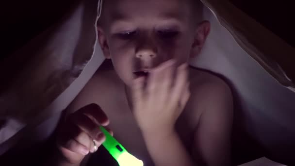 一个孩子晚上用手电筒在毯子下看书。轻的头发和蓝色眼睛的男孩 — 图库视频影像