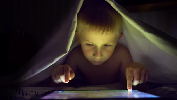 Young blond boy under covers playing on tablet a computer game. — Stock Video