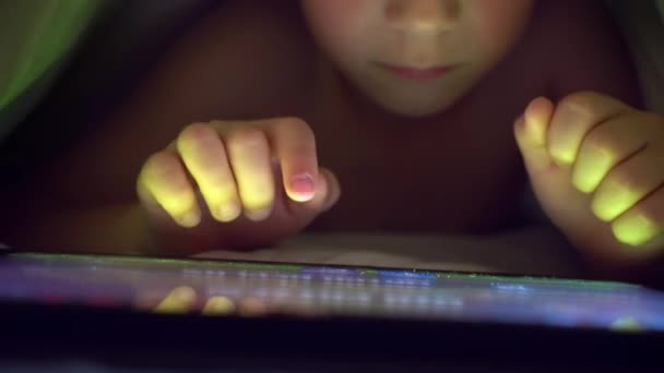 Kleiner blonder Junge spielt unter einer Decke auf dem Tablet ein Computerspiel. — Stockvideo