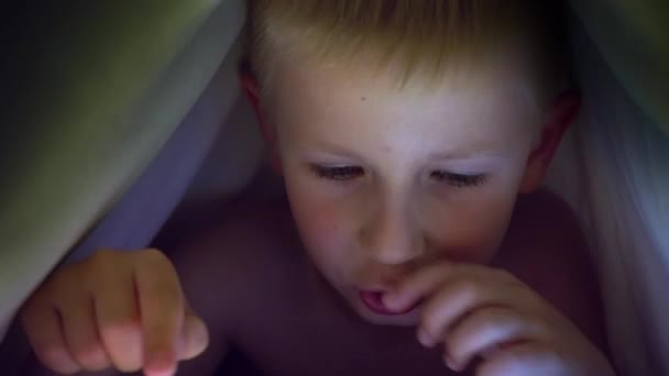 Kleiner blonder Junge spielt unter einer Decke auf dem Tablet ein Computerspiel. — Stockvideo