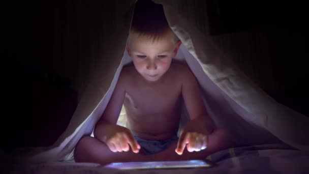 Young blond boy under covers playing on tablet a computer game. — Stock Video
